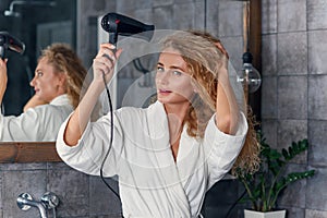 Close up of appealing smiling young lady in white dressing gown posing on camera while drying her curly hair using hair