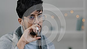 Close-up arabian indian bearded guy with glasses lonely sad sick ill man wrapped in blanket spraying rhinitis spray anti