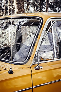 Close up artistic style view of old yellow vintage car front window wheel and antenna outdoors in summer.