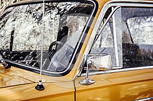 Close up artistic style view of old yellow vintage car front window wheel and antenna outdoors in summer.