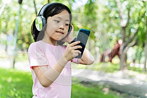 Close up of asian child girl with smartphone and listens to music with modern headphones wireless in park outdoors