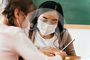 Close-up of Asian female teacher wearing a face mask in school building tutoring a primary student girl. Elementary