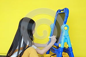 Close up asian little child girl with writing and drawing on empty blackboard over yellow background. Student and development