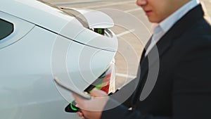 Close up of asian man in black business suit charging white luxury car and using modern smartphone. Electric vehicle