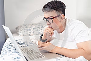Close up of Asian man wearing glasses using laptop computer working from home laying on bed, digital lifestyle social distancing,