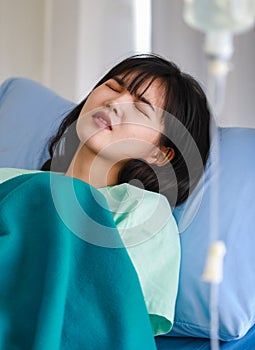 Close-up of an asian sick female patient covered with a blanket lying on hospital bed having intravenous drip saline solution,