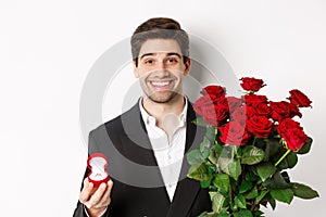 Close-up of attractive man in suit, holding bouquet of roses and engagement ring, making proposal, standing against