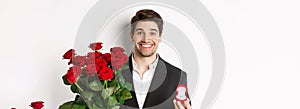 Close-up of attractive man in suit, holding bouquet of roses and engagement ring, making proposal, standing against