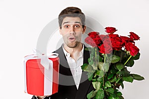 Close-up of attractive man in suit looking surprised, holding gift box and bouquet of roses, giving present for holiday