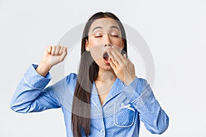 Close-up of attractive sleepy asian girl in blue pajama, waking-up in morning and stretching with closed eyes, yawning
