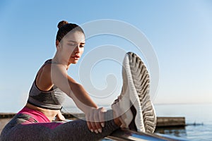 Close-up of attractive sportswoman holding leg on handrope, stretching on the seaside promenade. Female runner warming
