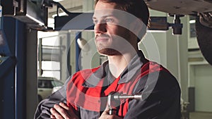 Close-up auto mechanic crossed hands and looking at camera while standing under lifting car in repair garage.