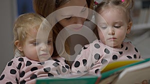 Close-up of baby twin sisters listening to mother reading book in the evening. Beautiful Caucasian daughters enjoying