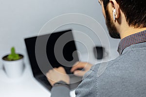 Close-up back view of businessman working at laptop, using wireless earphones.