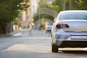 Close-up back view of new shiny expensive silver car moving along city street on blurred trees, cars and buildings background on