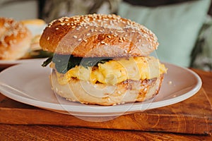 A close-up of a bagel burger with scrambled eggs, spinach, cheese, and avocado on a plate