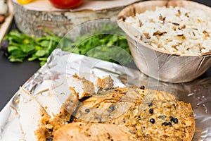 Close up of baked chicken breast on foil near plate of rice, veg