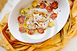 Close-up baked molten feta cheese with small tomatoes in olive oil with herbs and spices in ceramic orange baking dish and yellow