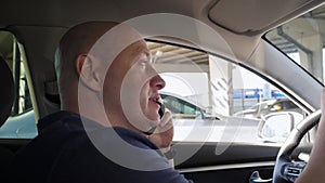 Close-up of a bald man talking on the phone while sitting in a car at the wheel.