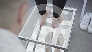 Close-up of barefoot female legs on transparent glass on medical examination. Shooting over shoulder of male orthopedist