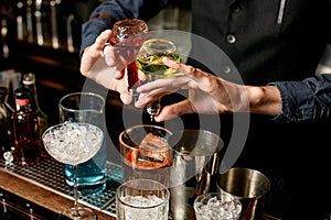 Close-up bartender holds bottles with liquor and adds it to glass with cocktail