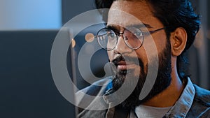 Close-up bearded male face reflection light from laptop monitor in glasses. Busy serious focused Arab Indian man looking