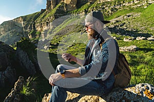 Close-up of a bearded man in jeans clothes in sunglasses and a cap with a backpack sitting at the foot of the epic rocks