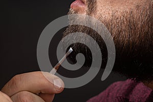 Close-up of bearded man pulling gray hair out of graying beard with tweezers. Concept of skin, hair care and anti-aging