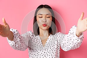 Close-up of beautiful asian woman beauty blogger, pucker lips and close eyes for kiss, taking selfie, standing over pink