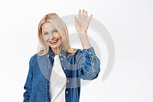 Close up of beautiful blond senior woman, happy lady saying hello, waving hand to say hi, standing in casual clothes