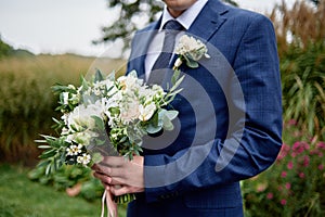 Close up of beautiful bridal bouquet of white and cream flowers and greenery in groom& x27;s hand outdoors, copy space.