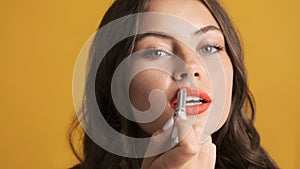 Close up beautiful brunette girl applying red lipstick on camera over colorful background