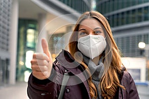 Close up of beautiful confident woman wearing protective mask KN95 FFP2 showing thumb up with modern city on background and