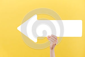 Close up of beautiful female hand holding big white paper arrow, isolated over yellow studio background wall with copy space