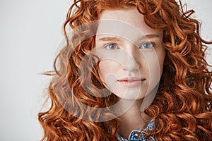 Close up of beautiful girl with curly red hair and freckles looking at camera over white background.