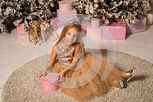 Close up. A beautiful little girl in an elegant golden dress sits on a soft carpet and smiles while holding a pink gift box