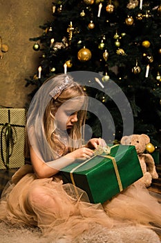 Close up. Beautiful little girl in a golden dress sits near an elegant Christmas tree and opens a green gift box