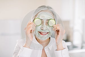 Close up of beautiful retired woman with facial mask on her face holding slices of fresh cucumber covering her eyes, on