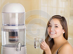 Close up of a beautiful smiling woman holding a glass of water, with a filter system of water purifier on a kitchen