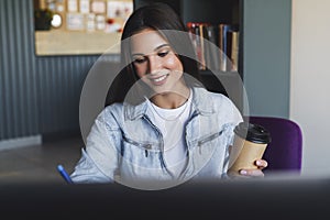 Close-up. Beautiful smiling young business woman sits in front of laptop, holds cup of coffee, makes notes in notebook