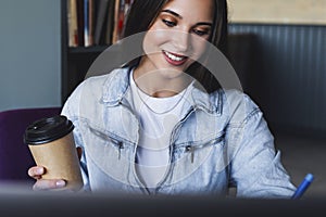 Close-up. Beautiful smiling young business woman sits in front of laptop, holds cup of coffee, makes notes in notebook
