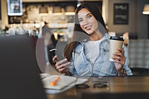 Close-up. Beautiful smiling young business woman sits in front of laptop, holds cup of coffee, uses smartphone for work