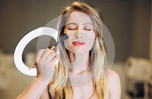 Close-up of a beautiful woman with blond hair and a powder brush near the face. To put powder on a beautiful face with a