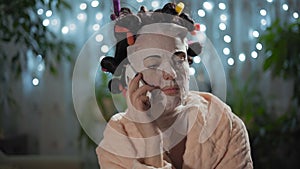 Close-Up, Beautiful Woman in a Rejuvenating Mask with Curlers Talking on the Phone Sitting in the Room