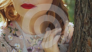 Close-up of beautiful woman's mouth with red lipstick, curling her hair standing near the tree.