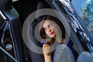 Close up of beautiful woman wearing a blue dress in a reclined seat and posing inside of a luxury black car on a