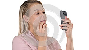 Beautiful young blonde woman which is painting her lips with red lipstick and looking in the phone on white background.