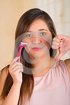 Close up of a beautiful young funny woman with an shaver on her face with a thumps down, in bath background