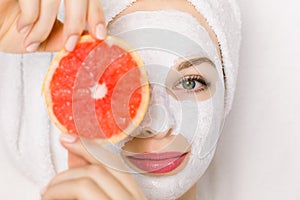 Close up of beautiful young smiling woman with towel on head, facial skin mask, holding grapefruit slice in front of her