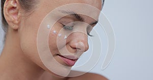 Close-up beautiful young woman applies face care cream on her cheek smiling for the camera on gray background. Skin care